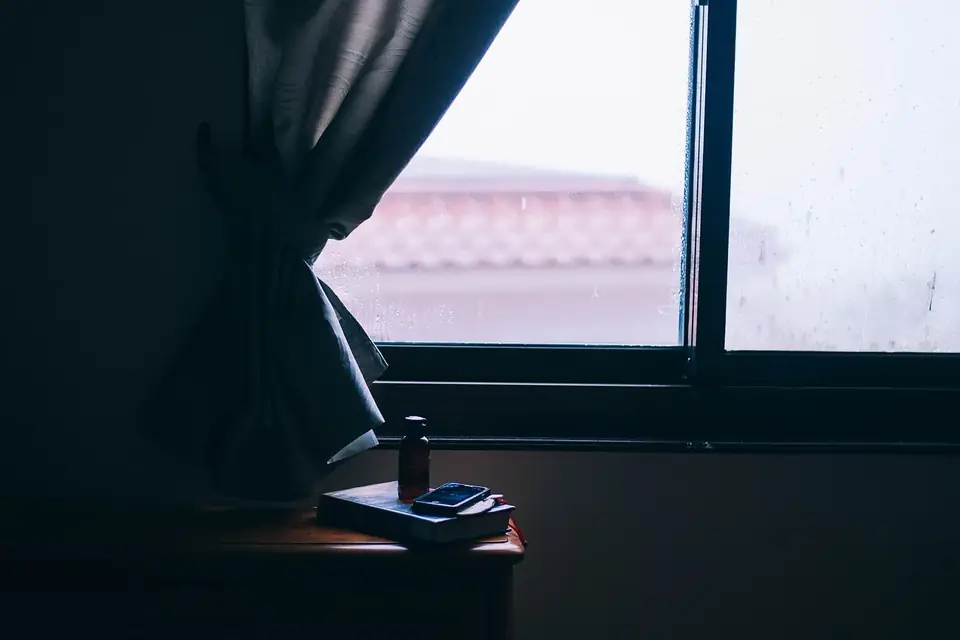 Rideau sombre devant une fenêtre embuée, un livre et un téléphone posés sur une petite table