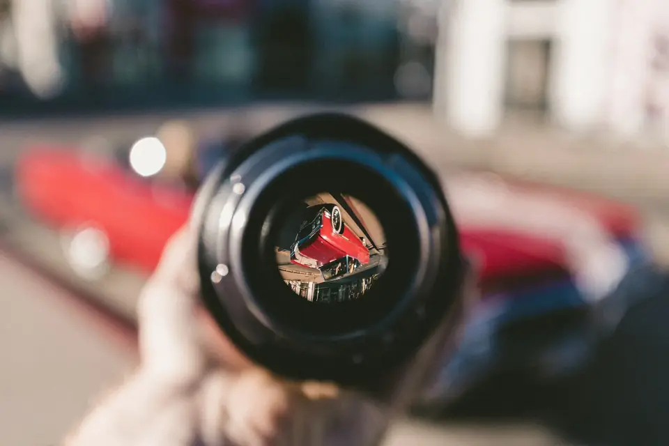 Main tenant un objectif photographique dans lequel se reflète une rue avec une voiture rouge