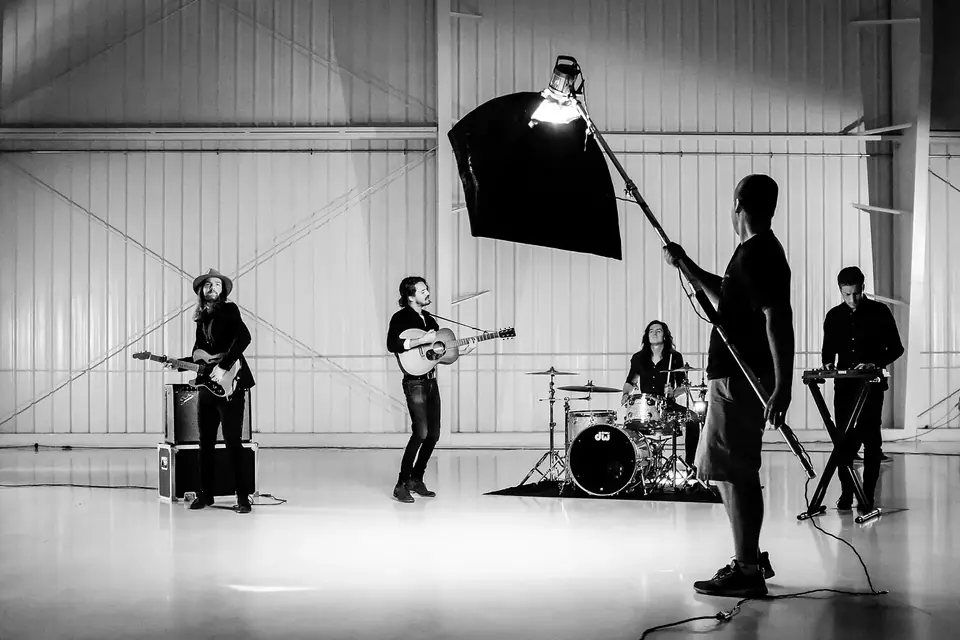Tournage en noir et blanc dans un hangar : un groupe de musiciens joue tandis qu'un technicien tient une boîte à lumière sur une perche